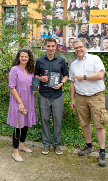 Geschenkübergabe zum 20 jährigen Jubiläum. 1 Frau und 2 Männer lächeln in die Kamera.