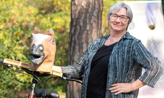 Geschäftsführerin von Sandstein Kultur lächelt in die Kamera. Sie hält ein Fahrrad mit Pferdemaske am Lenker fest.