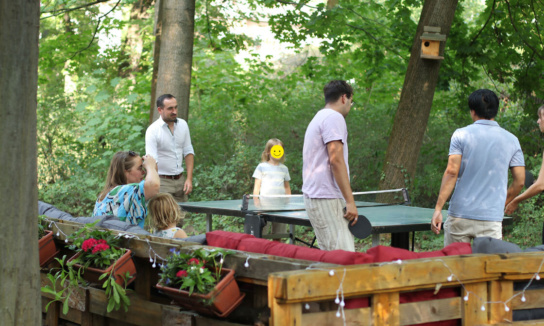 5 Personen spielen Tischtennis. 1 Frau und 1 Kind sitzen davor.