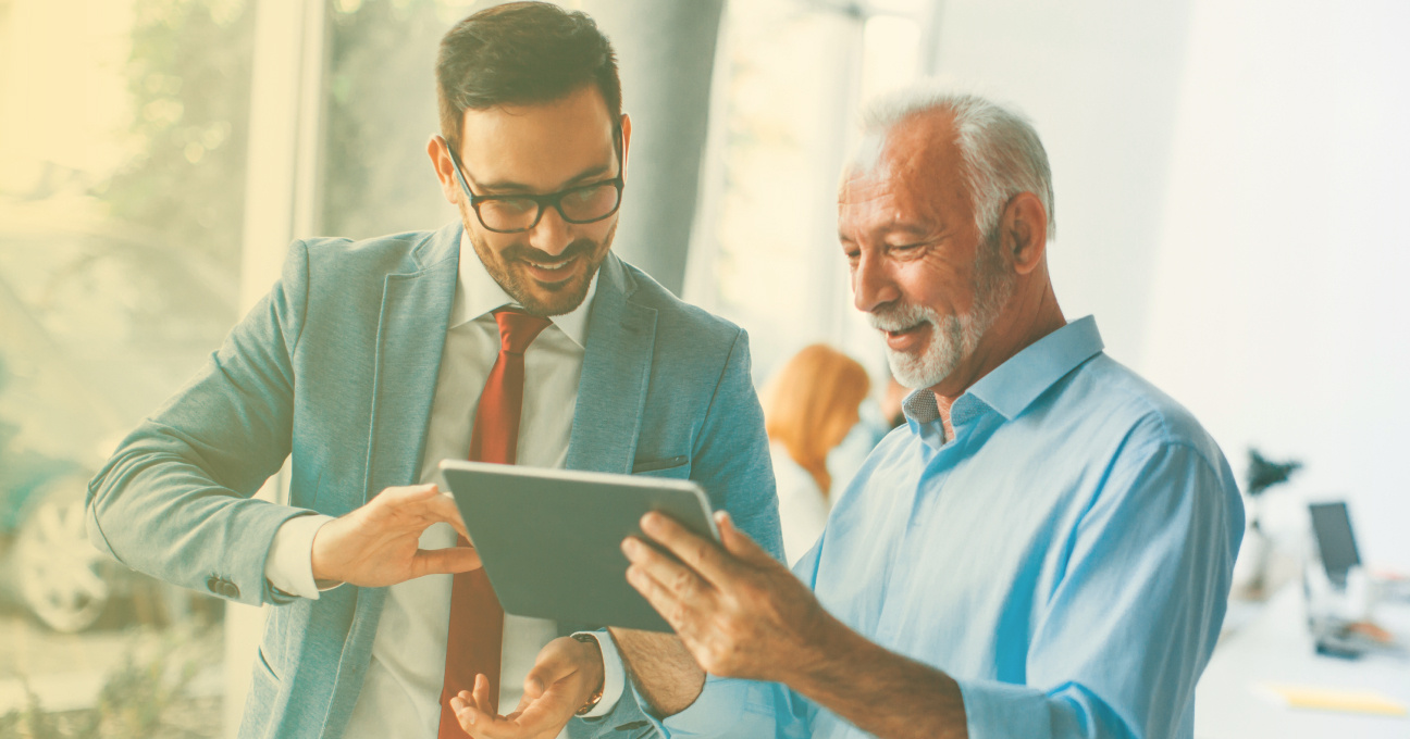Foto: Zwei Männer schauen zusammen auf ein Tablet. Ein Mann ist jung und trägt einen Anzug. Der andere Mann ist älter und hat weiße Haare und einen Bart. Beide Männer lachen und sprechen miteinander.