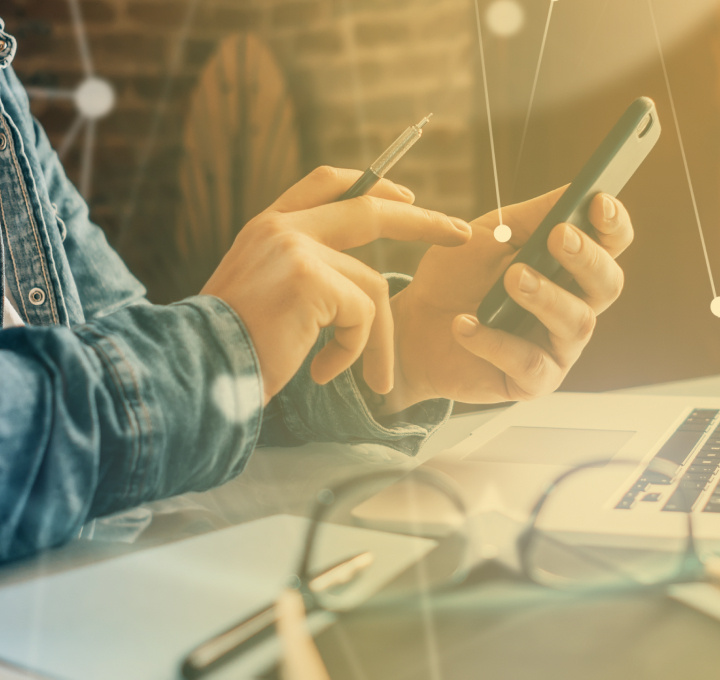 Foto: Eine Person sitzt an einem Schreibtisch. Die Person hält ein Handy in der Hand und tippt darauf. Neben dem Handy liegen ein Laptop, eine Brille und ein Stift.