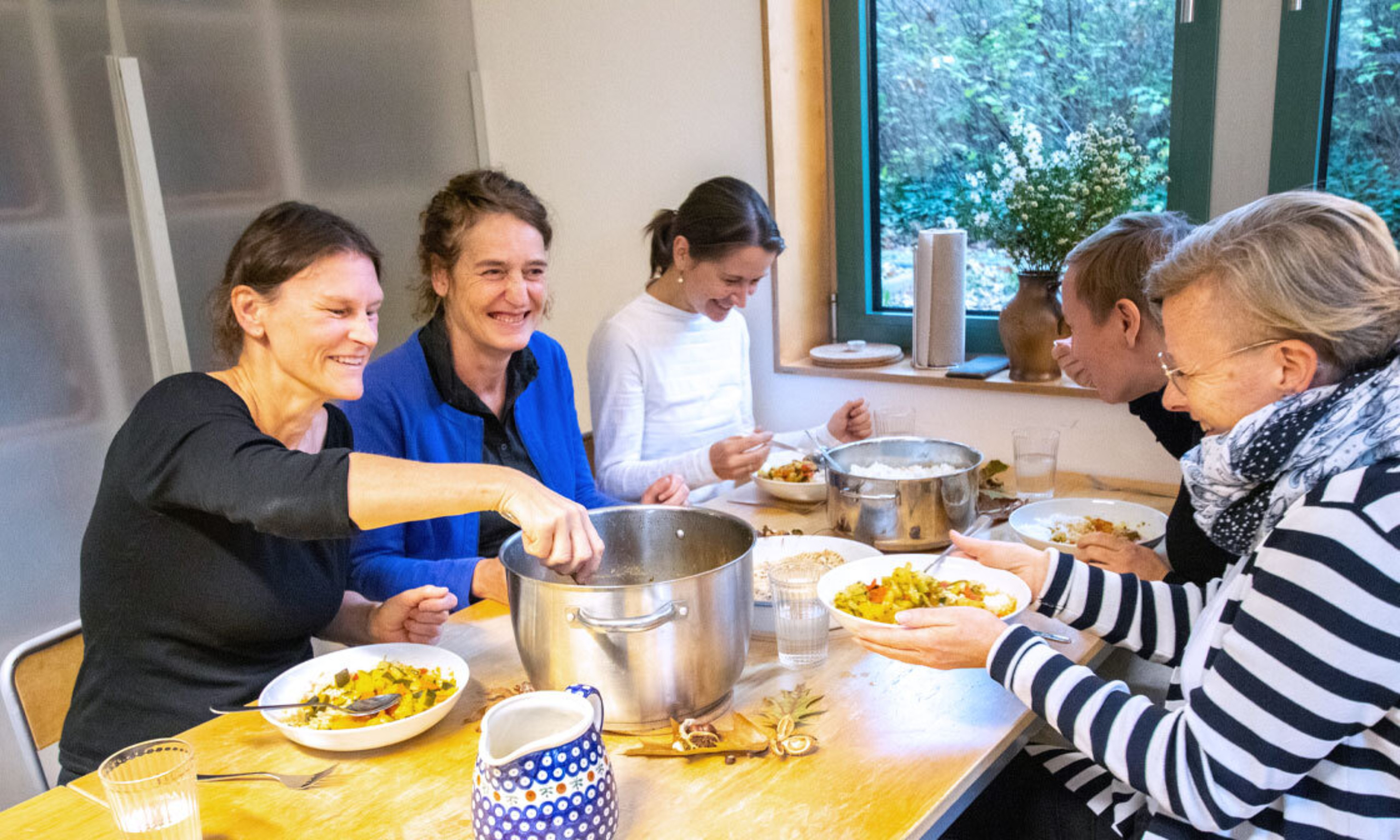 5 Frauen sitzen gemeinsam am Tisch. Auf dem Tisch steht ein großer Topf mit Ratatouille.