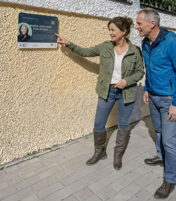 ein Mann und eine Frau zeigen auf eine Plakette an einer Mauer