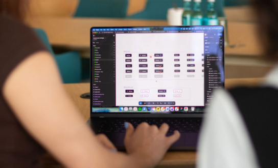 Blick auf ein Display eines Laptops. Darauf verschiedene Modi von Buttons zu sehen. Im Vordergrund unscharfe Ansicht zweier Personen.