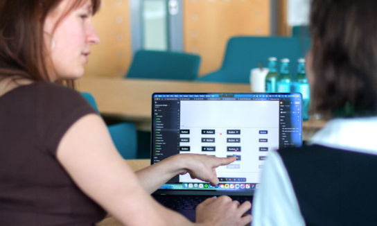 Blick auf ein Display eines Laptops. Darauf verschiedene Modi von Buttons zu sehen. Im Vordergrund zwei Personen, die darüber sprechen.