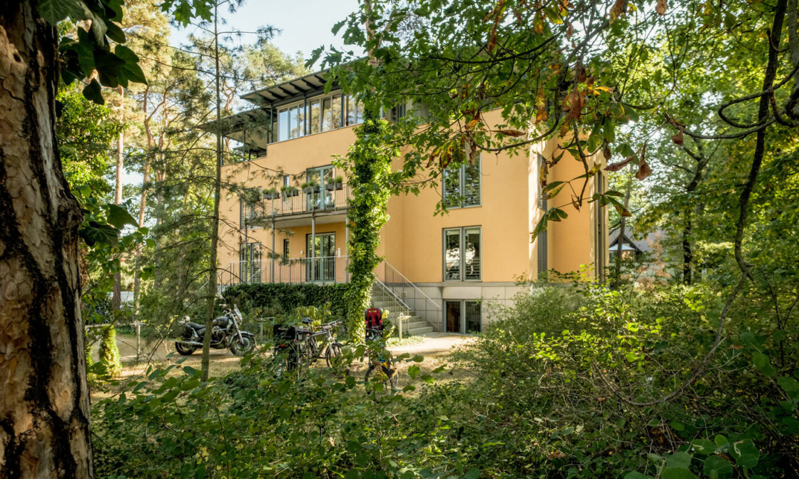 Das Gebäude der Firma Sandstein zeigt ein gelbes Haus mit vier Stockwerken umgeben von grünen Bäumen