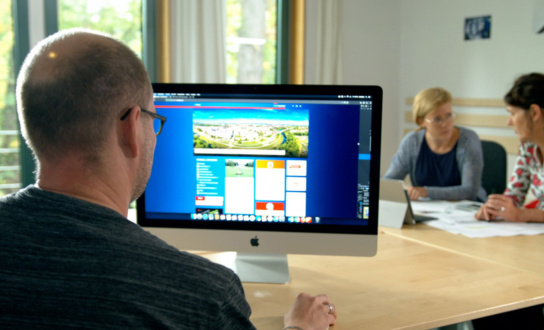 Mit dem Rücken zum Bild sitz ein Mann an einem Monitor. Dahinter am Tisch sitzen zwei Frauen, die miteinander sprechen.