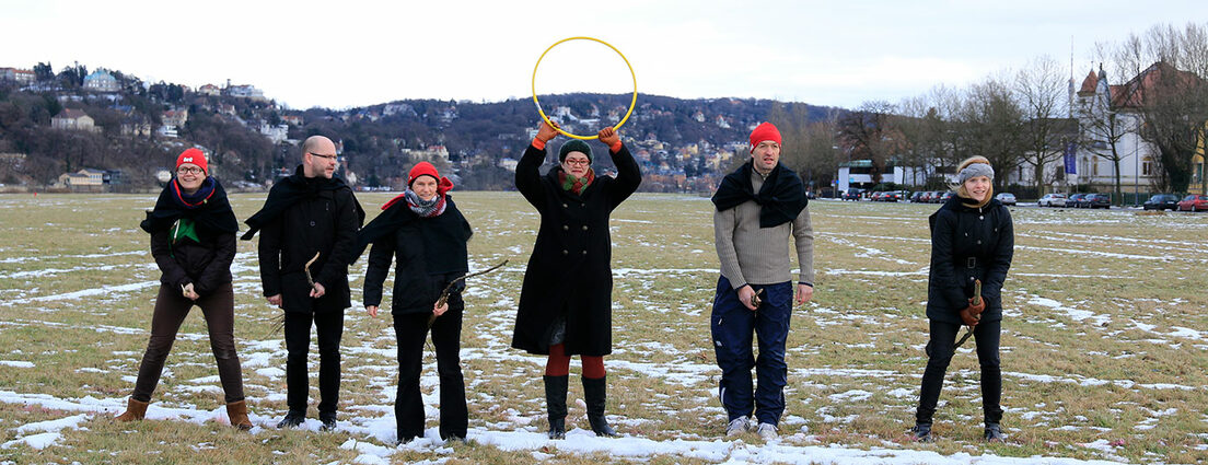 Foto: Personengruppe in Mänteln auf einer Wiese mit Stöcken und Reifen